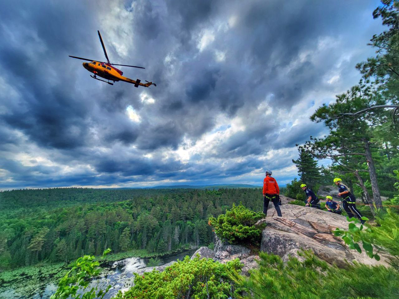 helicopter hovering in front of an ominous sky with four paramedics looking up from a rock cliff