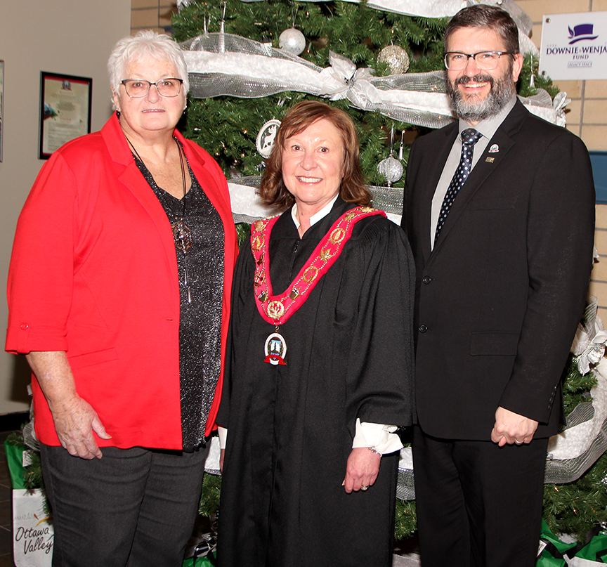 three people standing in front of a Christmas tree, warden in the middle wearing a robe and chain of office