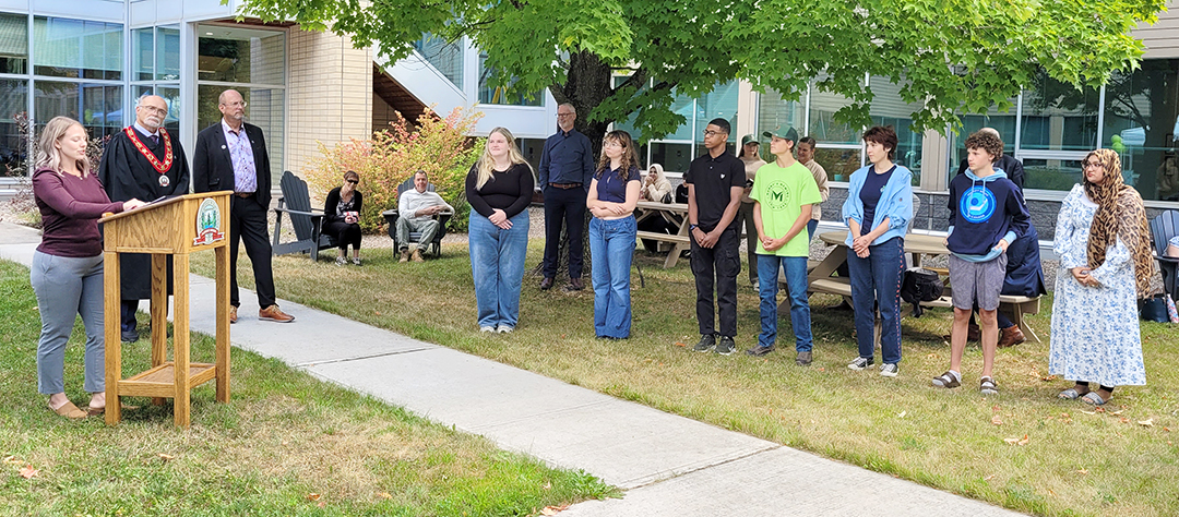 person at a podium with two people to the right and a row of students looking on in an outside setting