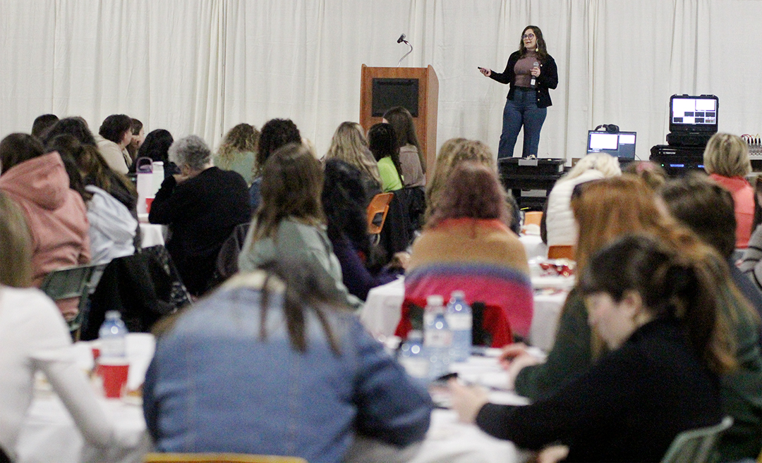 Woman in focus speaking on a stage with the blurred crowd in the foreground 